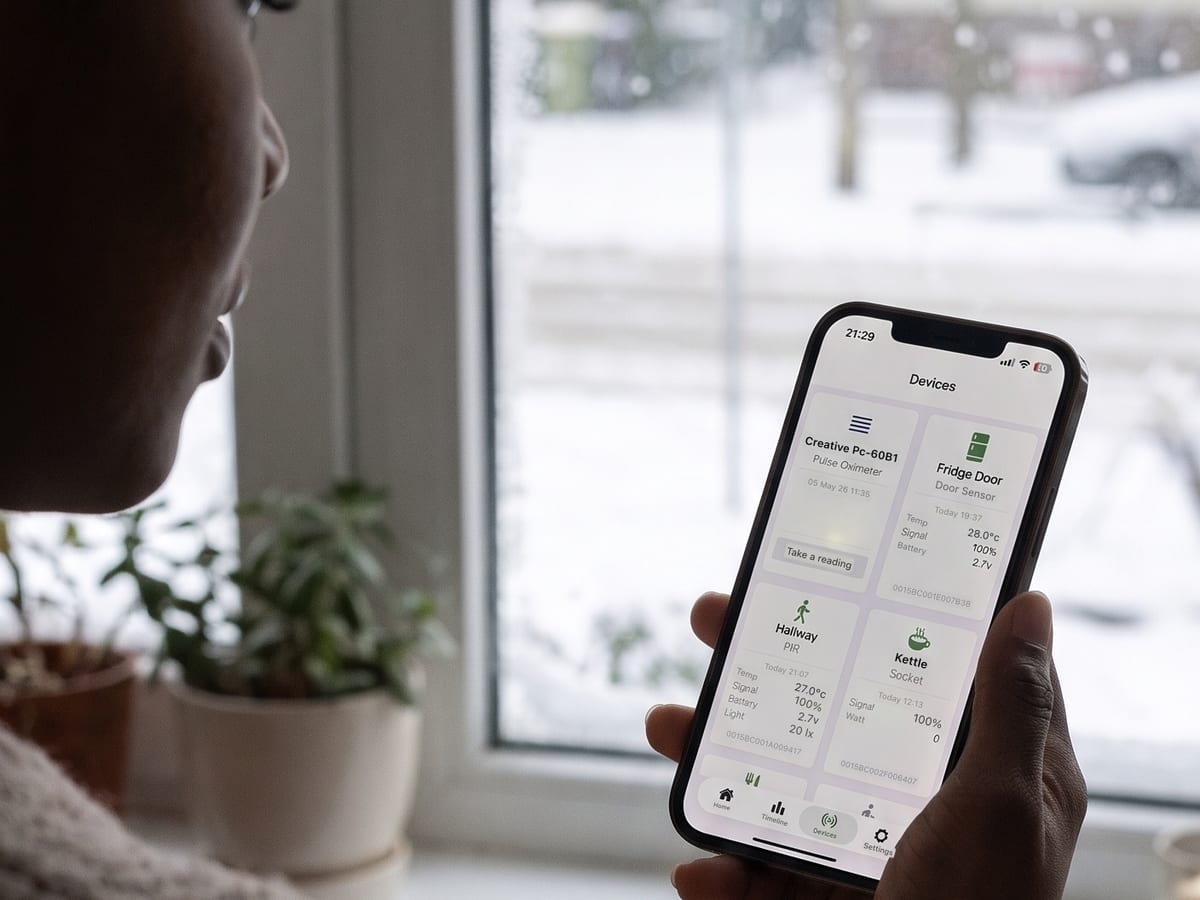 Someone by a snowy window checking the Howz app devices screen, which shows the sensors in the home with their temperature, signal and battery readings