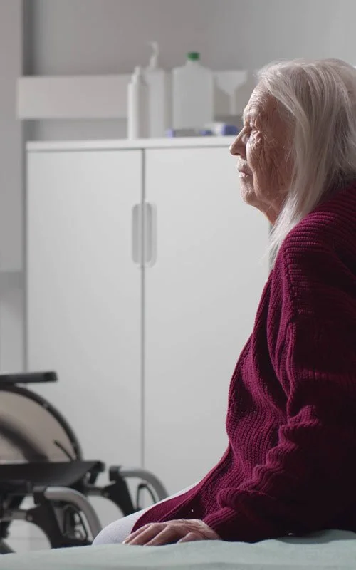 An elderly lady sitting on a hospital bed with a wheelchair in the room